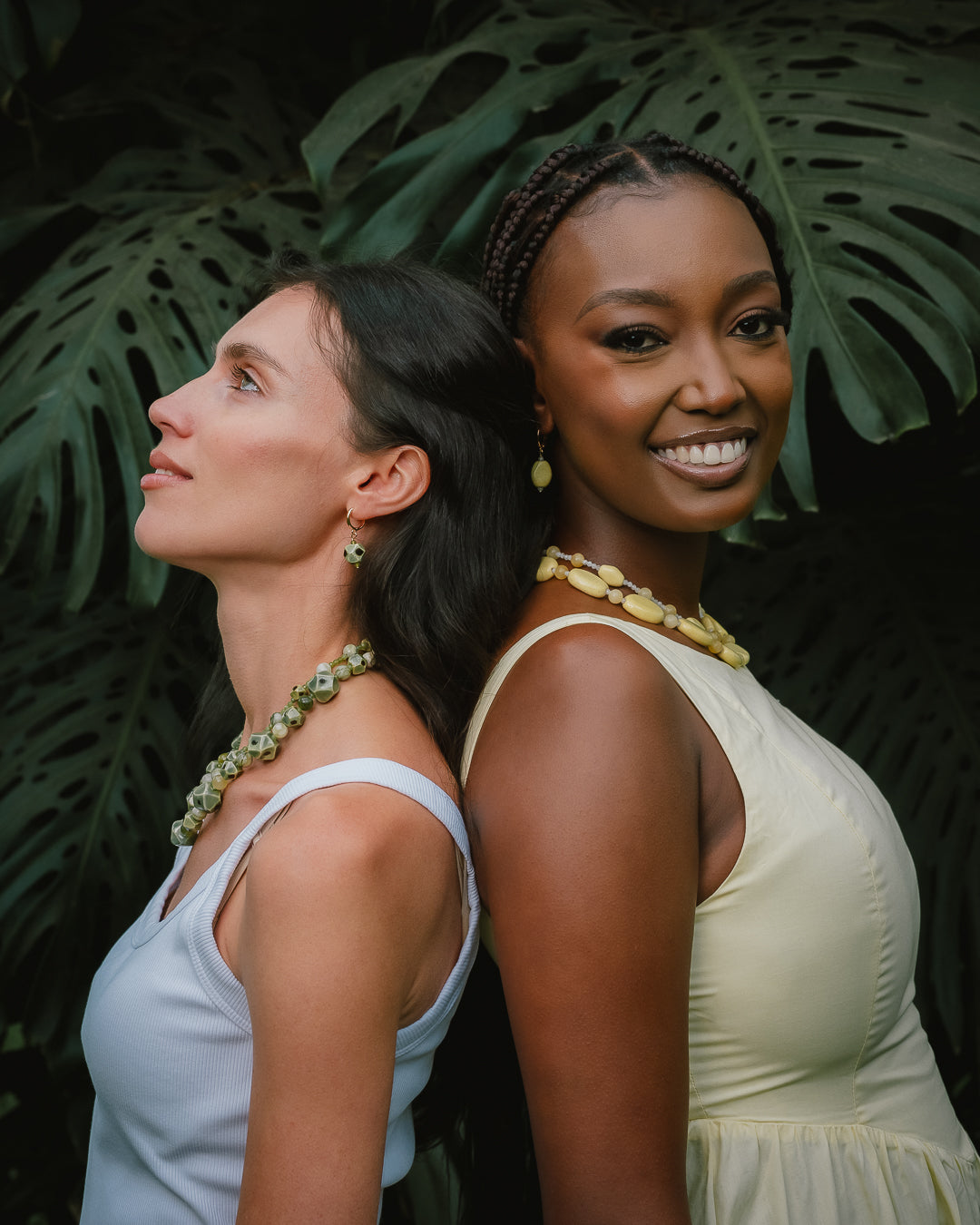 Two women standing back-to-back in front of large green tropical leaves, wearing matching light-colored dresses and matching beaded jewelry. The woman on the left wears a pale blue top and green beads, while the woman on the right wears a cream dress and yellow-green beads.